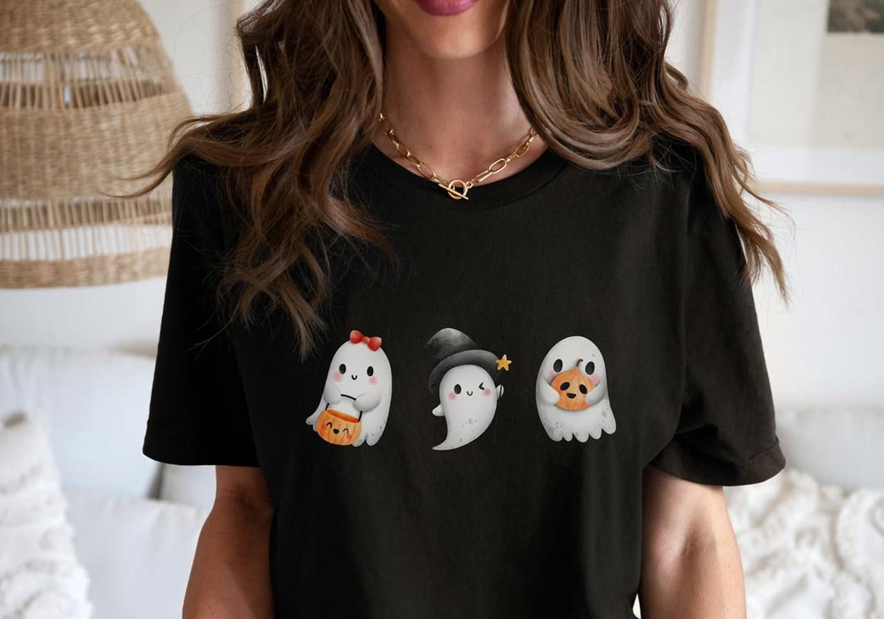 A woman wears a black t-shirt featuring three ghost illustrations. Each ghost has an accessory: a bow, a witch hat, and a pumpkin mask.
